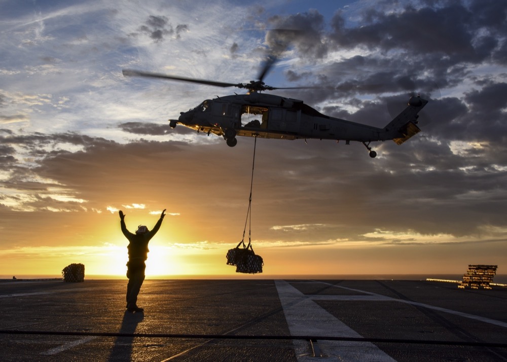 Helicopter lifting a slung load at sunset while a crewman signals